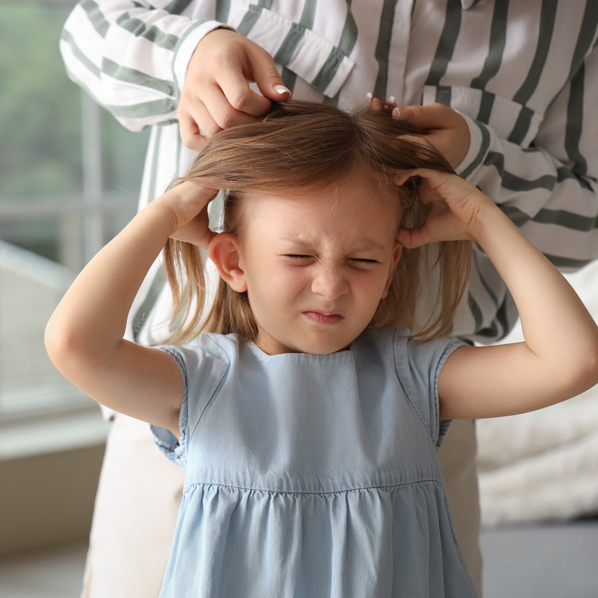 A litte girl holds her hair while her mother stands out of frame behind her checking her hair for lice.