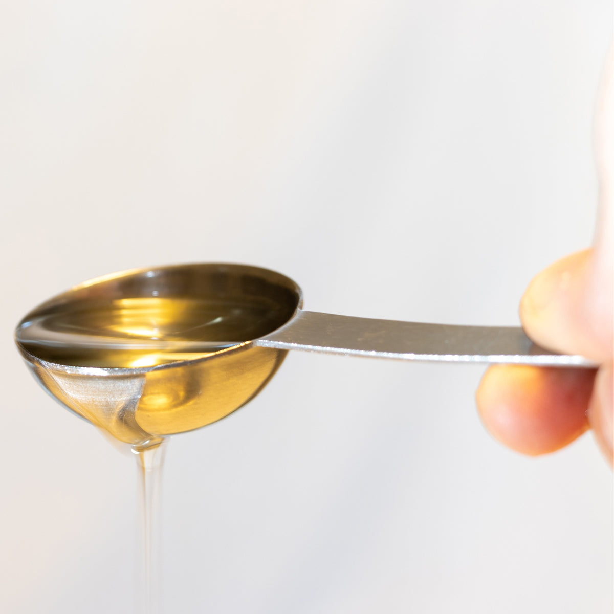 Hand holding a metal scoop with oil dripping onto a white background